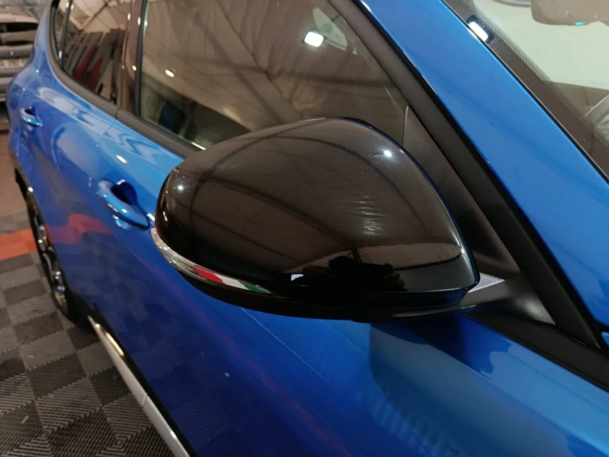 Gros plan sur le rétroviseur noir côté gauche de l'Alfa Romeo Tonale bleu, avec reflet du plafond de l'atelier.