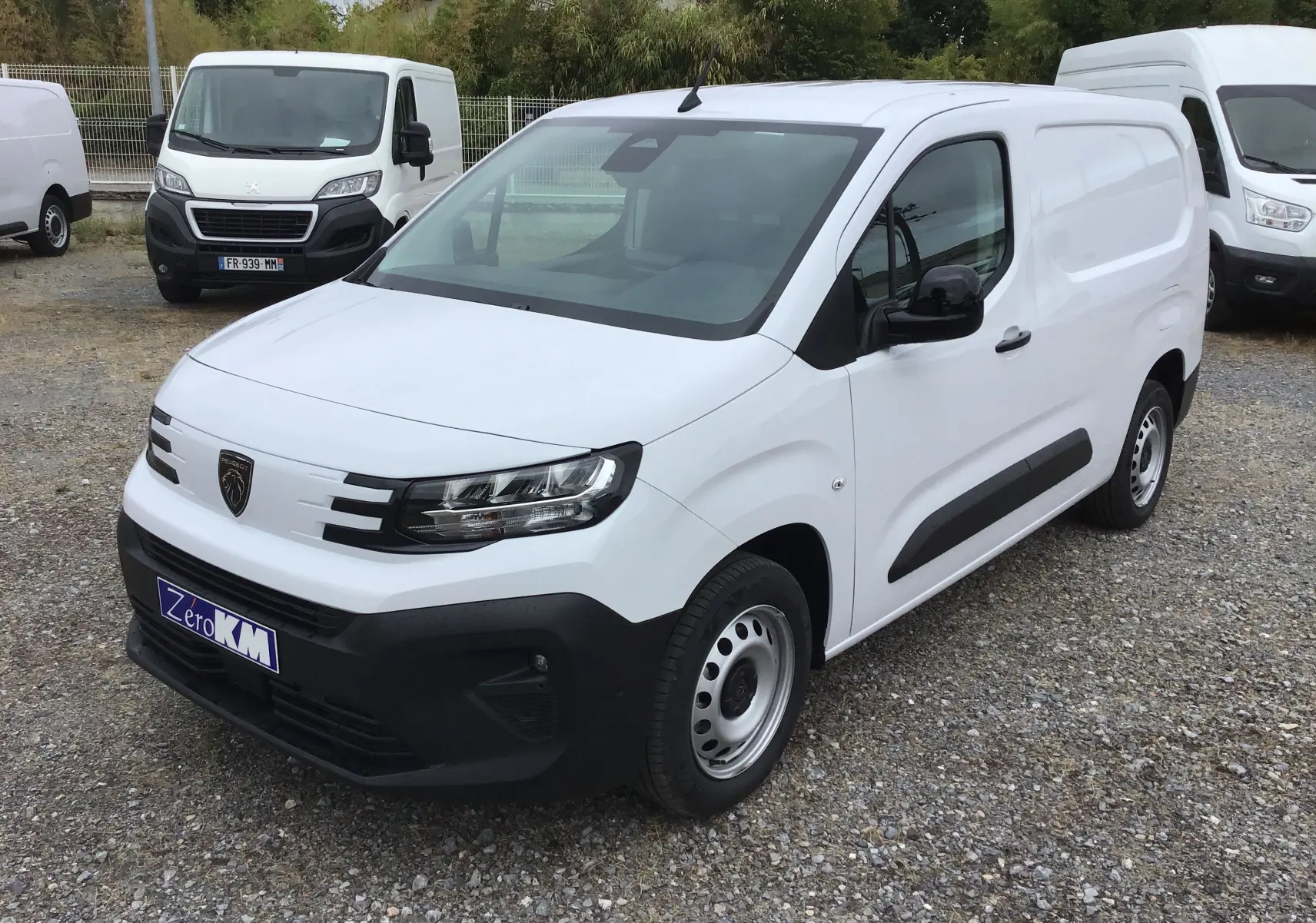 Peugeot Partner blanc en vue 3/4 avant droit, avec calandre noire et jantes acier, stationné sur un sol gravillonné.