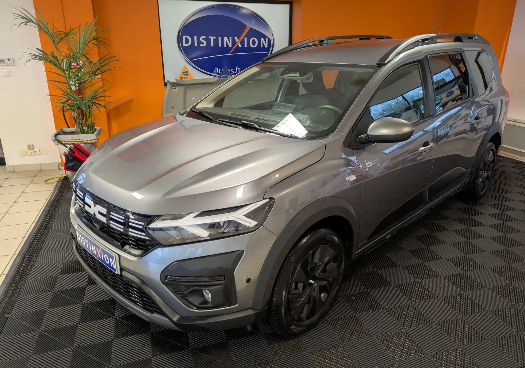 Vue 3/4 avant droit d’un Dacia Jogger gris schiste métal 2025 avec barres de toit et jantes noires en showroom.