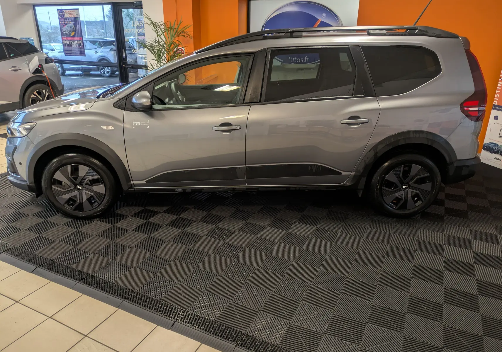 Vue de profil côté gauche d'un Dacia Jogger gris schiste métal, avec jantes noires et intérieur visible en showroom.
