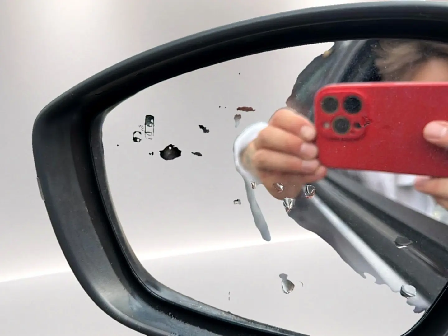 Rétroviseur droit d’un Opel Mokka gris 2025 avec traces d’eau et reflet d’une main tenant un smartphone rouge.