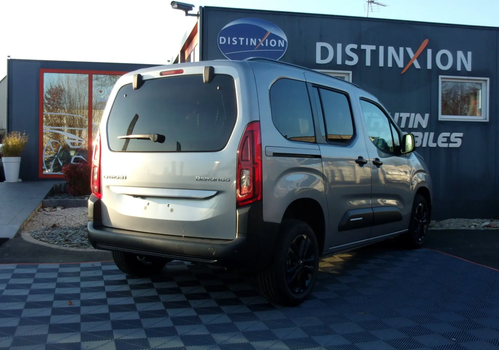 Vue 3/4 arrière droite d'un Citroën Berlingo gris Artense 2025, avec vitres teintées et jantes noires, stationné devant un bâtiment.