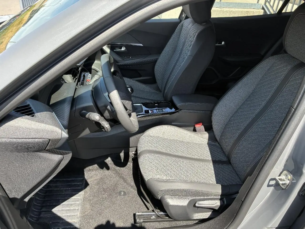 Vue intérieure côté conducteur du Peugeot 2008 1,2 100 CV Allure 2024, sièges tissu gris avec détails noirs, volant et console visibles.