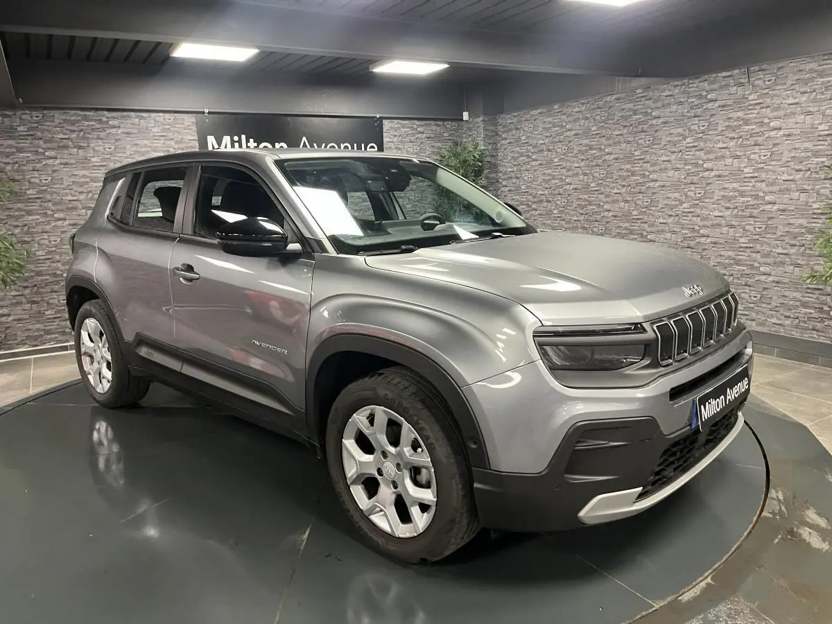 Jeep Avenger gris métallisé en 3/4 avant droit, avec calandre distinctive et coques de rétroviseurs noires brillantes.
