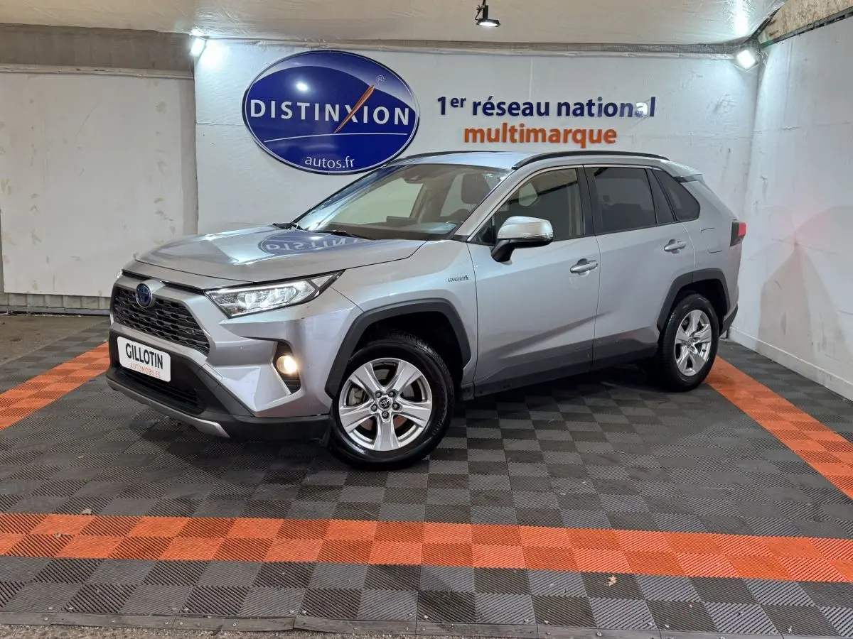 Toyota RAV4 hybride gris clair en 3/4 avant droit, avec phares allumés et jantes alliage visibles