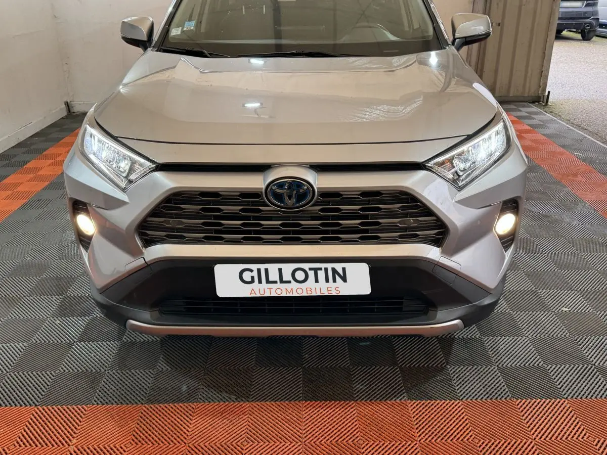 Vue frontale d'un Toyota RAV4 gris clair avec phares et antibrouillards allumés, calandre noire hexagonale.