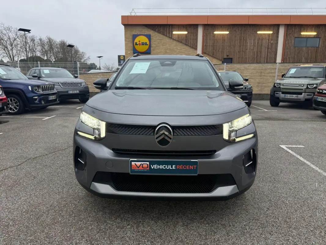 Vue frontale d'une Citroën C3 gris 2025 avec phares allumés et barres de toit visibles en arrière-plan.