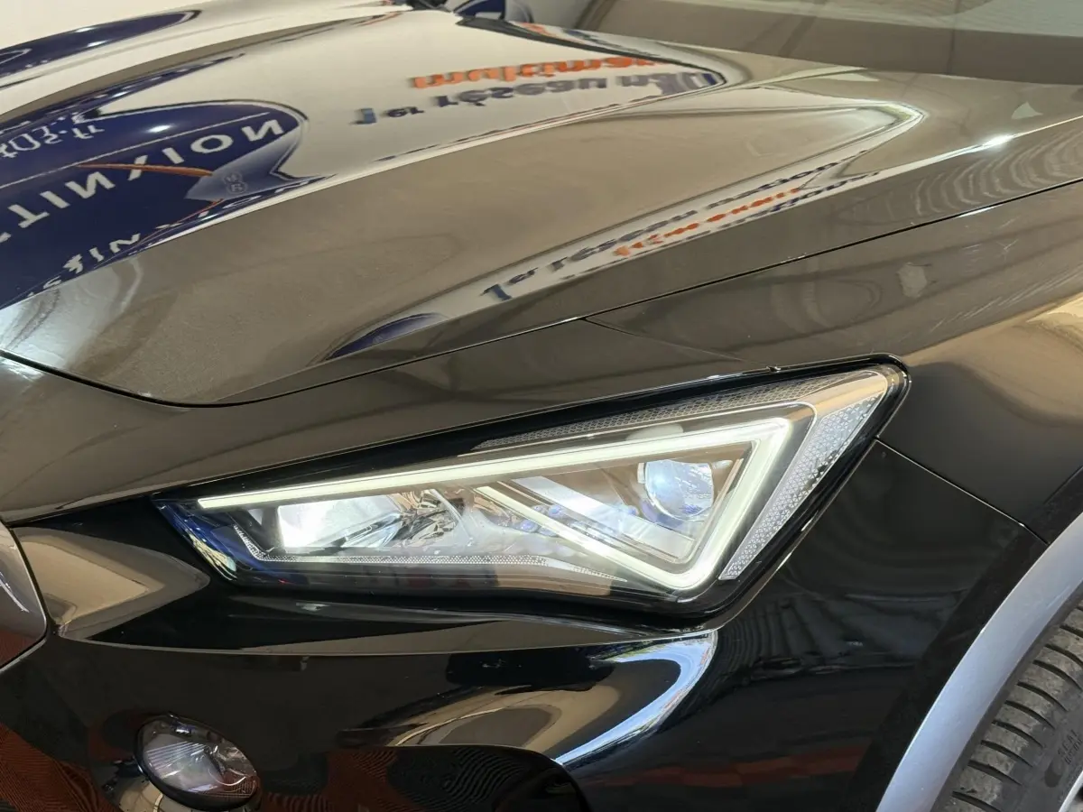 Gros plan sur le phare avant droit LED d'une CUPRA Formentor noire brillante avec capot lisse et reflet d'enseigne.