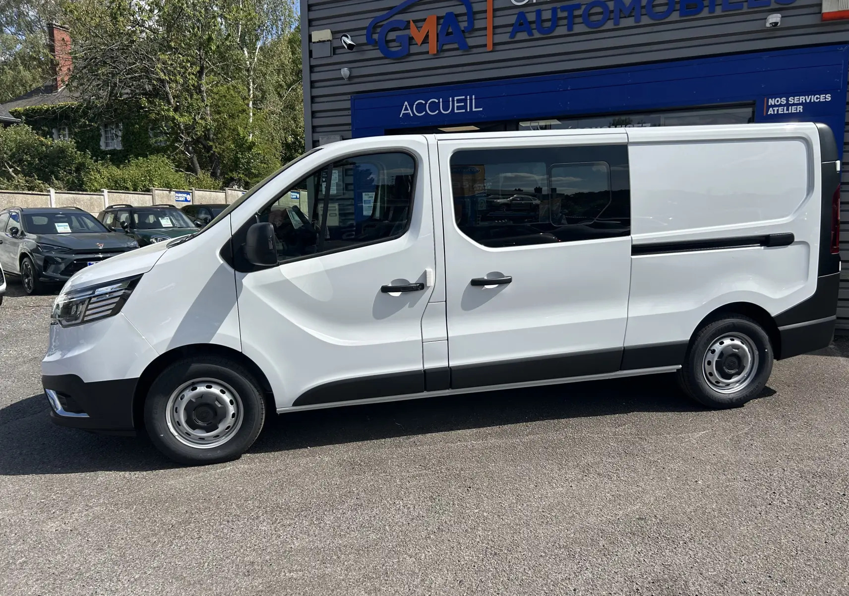 Profil droit d’un Renault Trafic cabine approfondie blanc, avec portes coulissantes et vitres teintées arrière, stationné devant un garage.