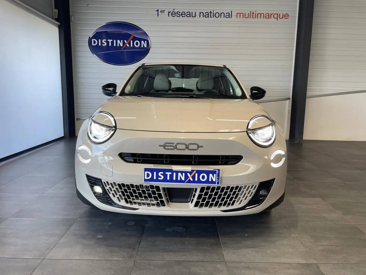 Vue frontale d'une FIAT 600 beige métallisé avec phares LED allumés et calandre ajourée, en showroom.