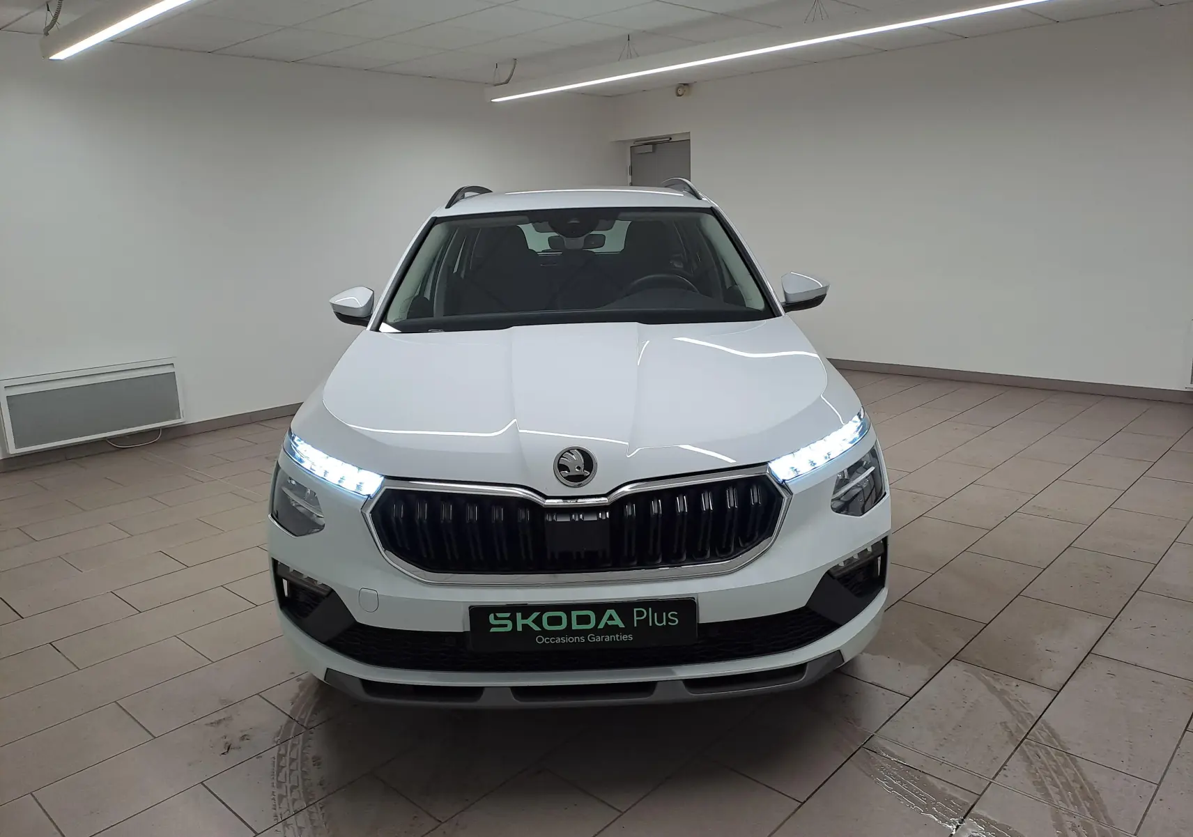 Vue frontale du Skoda Kamiq blanc Lune 2025 avec phares allumés et calandre noire dans un showroom.