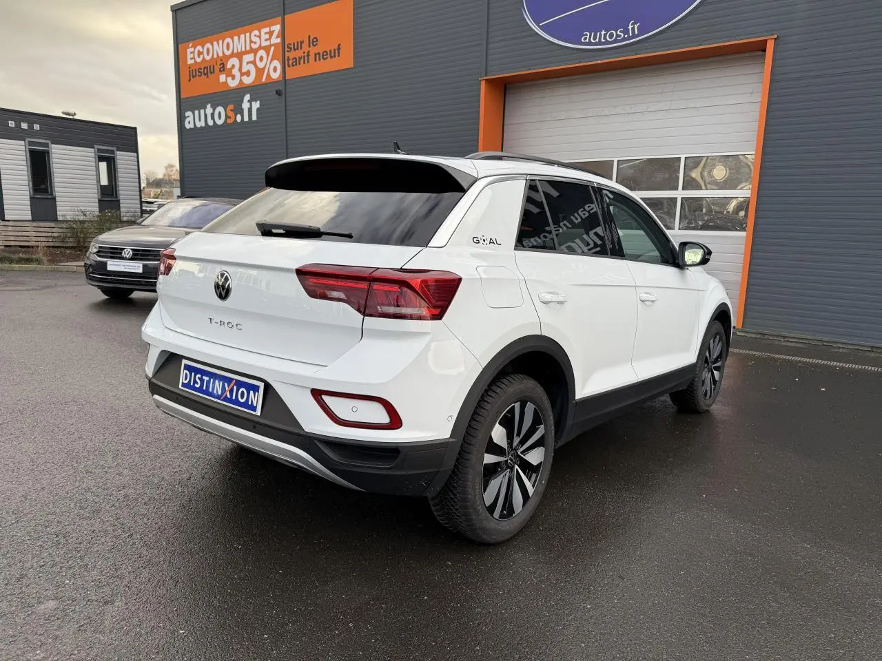 Vue 3/4 arrière droite d'un Volkswagen T-Roc blanc pur avec jantes alliage Zürich, stationné devant un garage autos.fr.