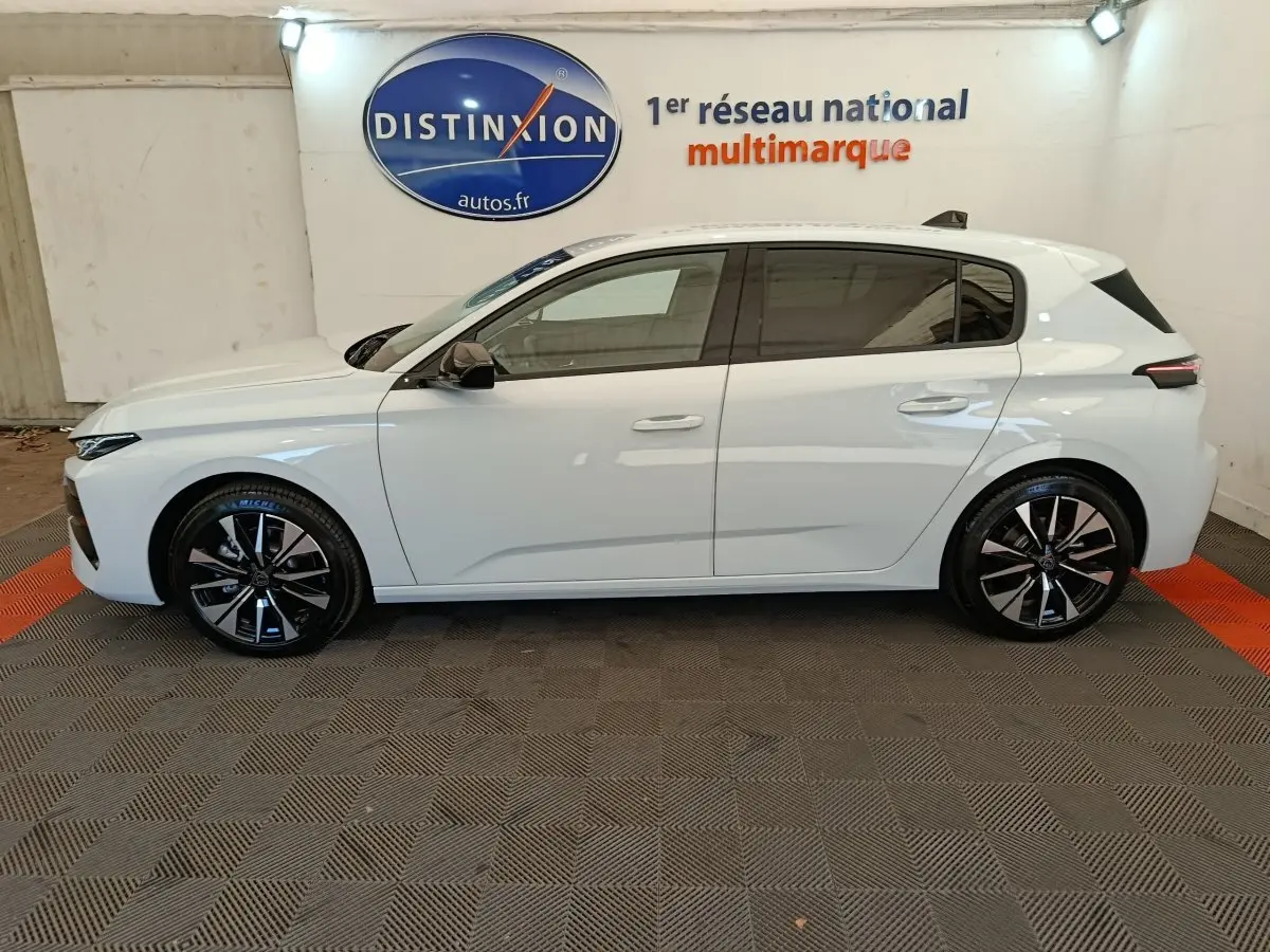 Vue de profil côté gauche d'une Peugeot 308 blanche 2026 dans un showroom avec jantes noires et logo Distinxion en arrière-plan.
