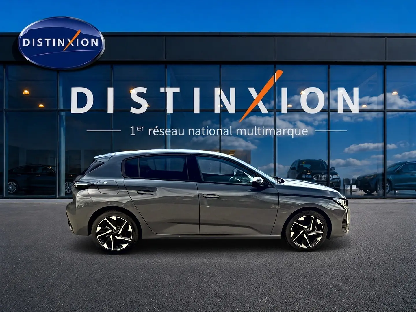 Profil droit d'une Peugeot 308 gris selenium 2025 garée devant une vitrine avec logo Distinxion, jantes alu noires et argentées.