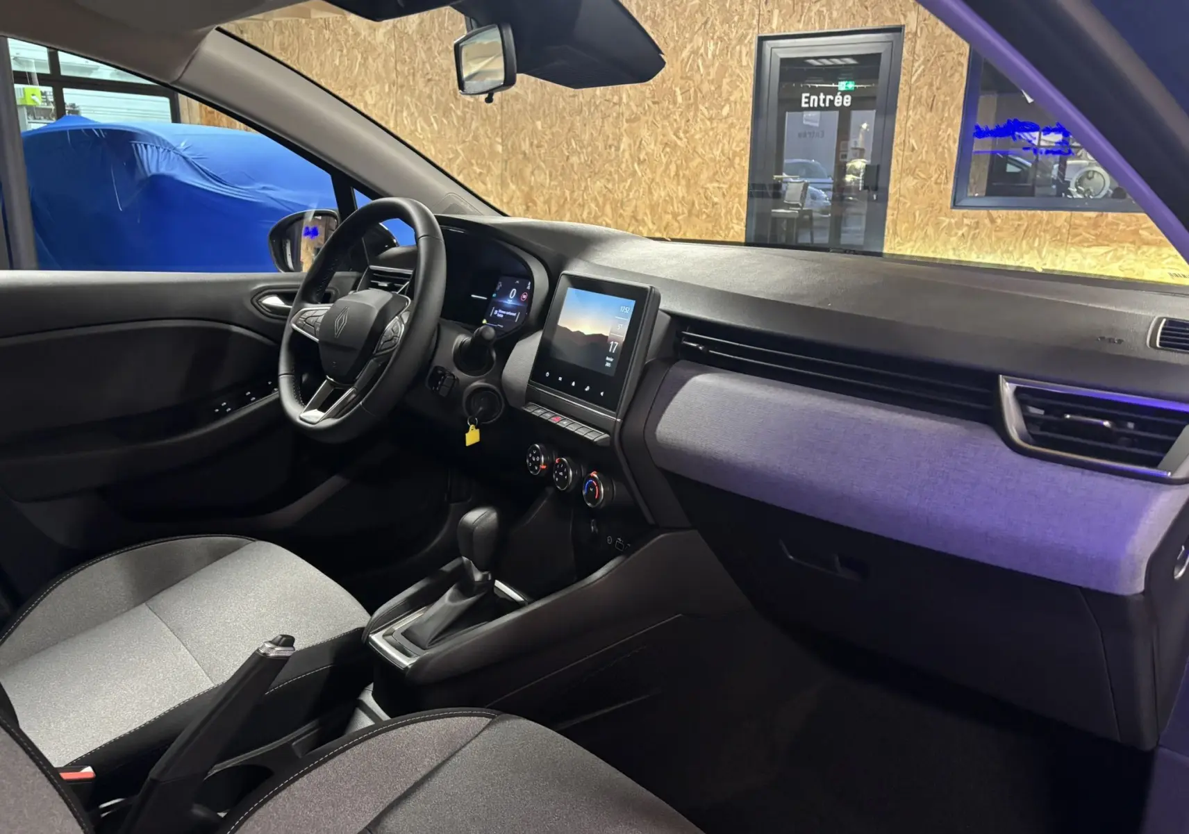 Intérieur de la Renault Clio 2024 vue côté conducteur, tableau de bord moderne avec écran tactile et sellerie tissu gris-noir.