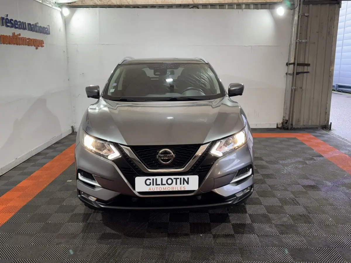 Vue frontale d'un Nissan Qashqai gris foncé avec phares allumés dans un garage éclairé.