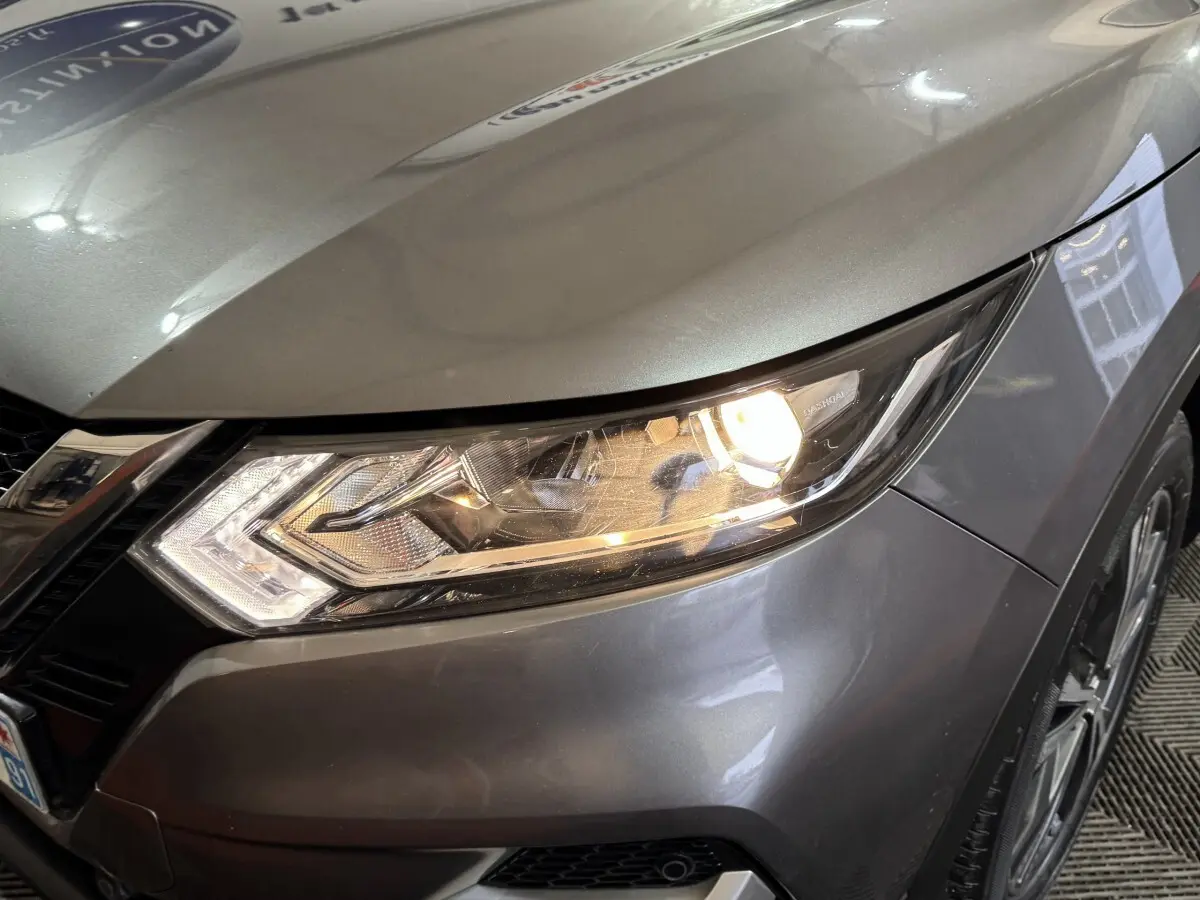 Gros plan sur le phare avant droit allumé d’un Nissan Qashqai gris foncé, avec partie du capot et de la roue visible.