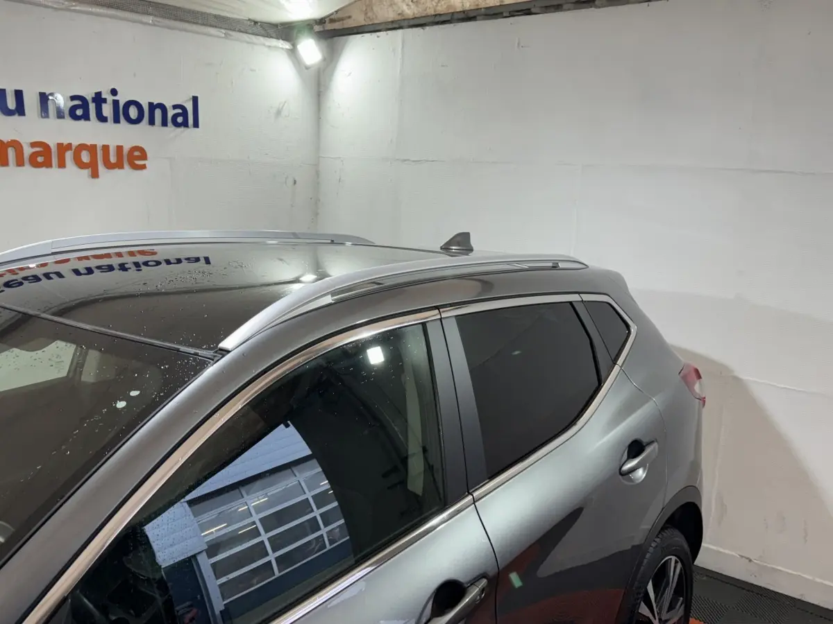 Vue latérale gauche d'un Nissan Qashqai gris foncé avec toit noir et barres de toit aluminium en intérieur showroom.