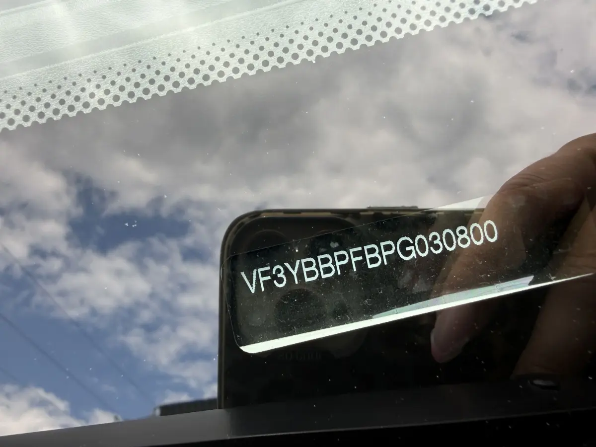Vue rapprochée du pare-brise avec numéro VIN visible, reflet du ciel et main tenant un smartphone.