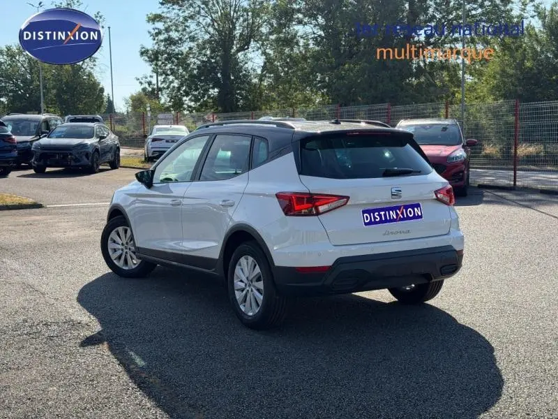 Vue 3/4 arrière droite d'une SEAT Arona blanc Candy avec toit noir Minuit, jantes alu et barres de toit visibles.