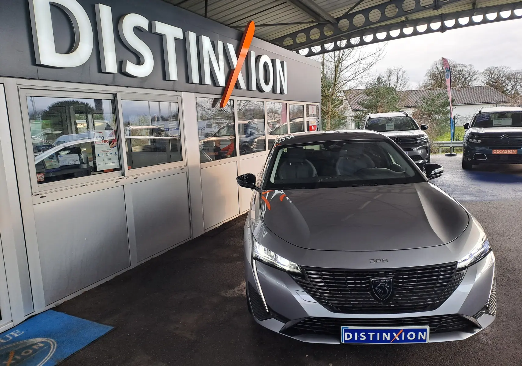 Peugeot 308 gris Artense vue de face sous un auvent avec logo Distinxion en arrière-plan.