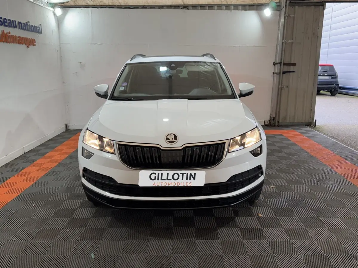Vue frontale d'un Skoda Karoq blanc avec calandre noire et phares allumés dans un garage éclairé.