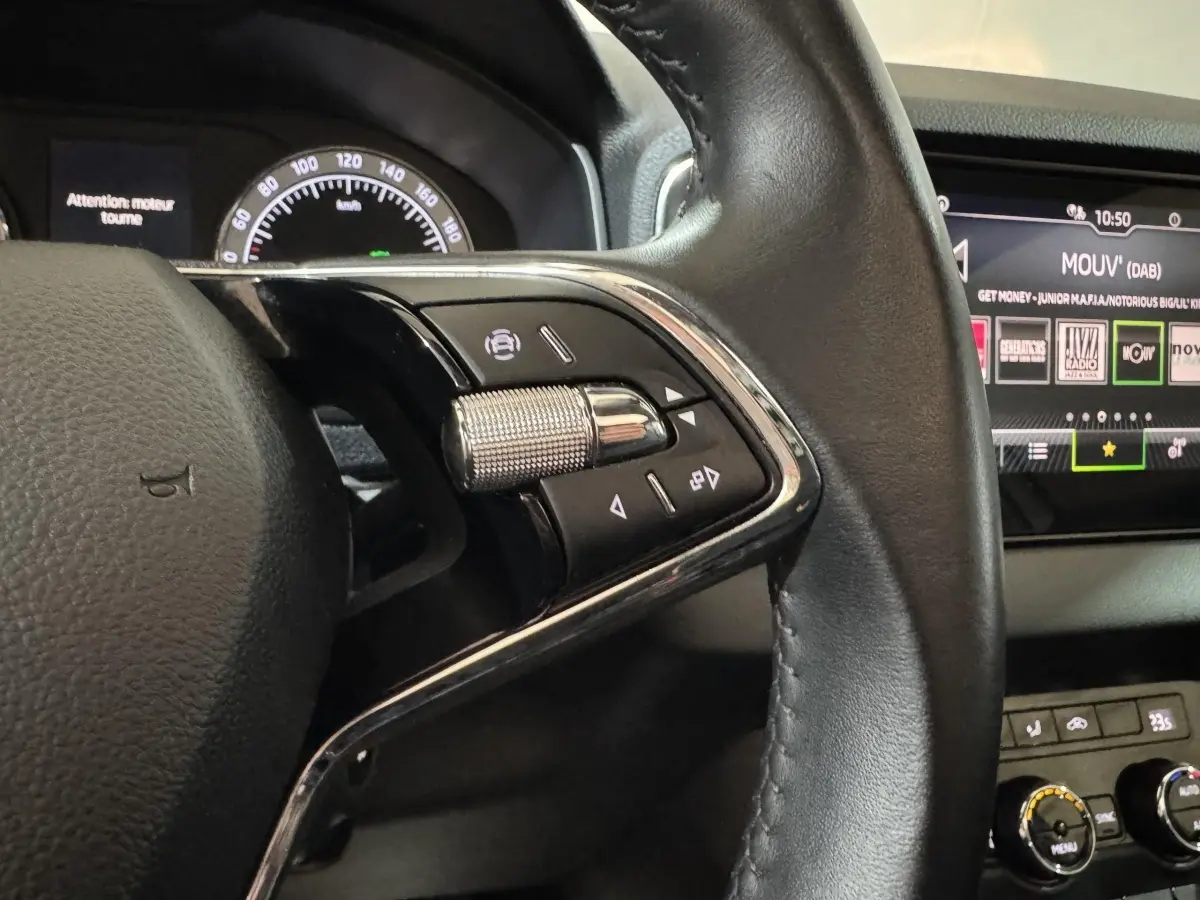 Gros plan sur la partie gauche du volant cuir noir du Skoda Karoq blanc, avec commandes multifonctions et écran tactile en arrière-plan.