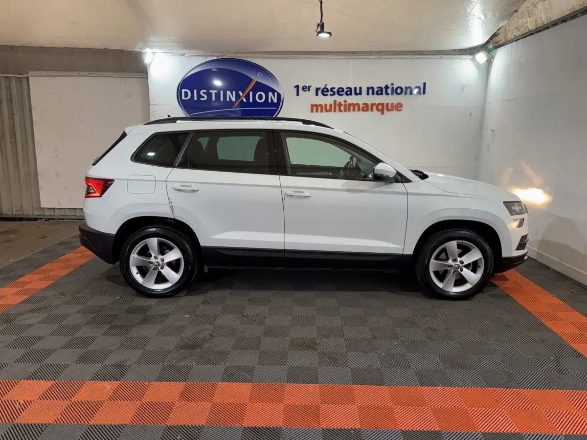 Profil droit d'un SUV SKODA KAROQ blanc 2021, avec jantes alliage et rails de toit noirs, en intérieur showroom.