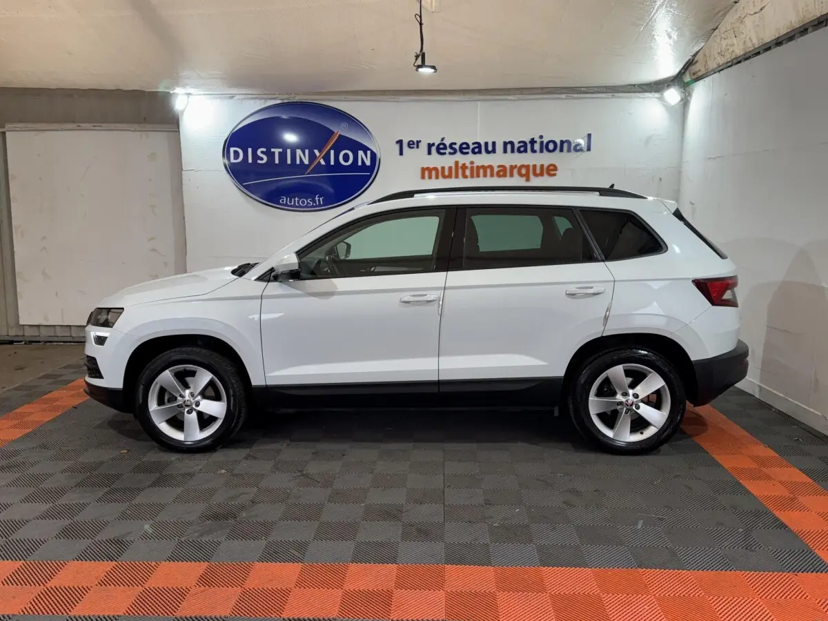 Vue latérale droite d'un SKODA KAROQ blanc 2021 avec barres de toit noires dans un showroom.