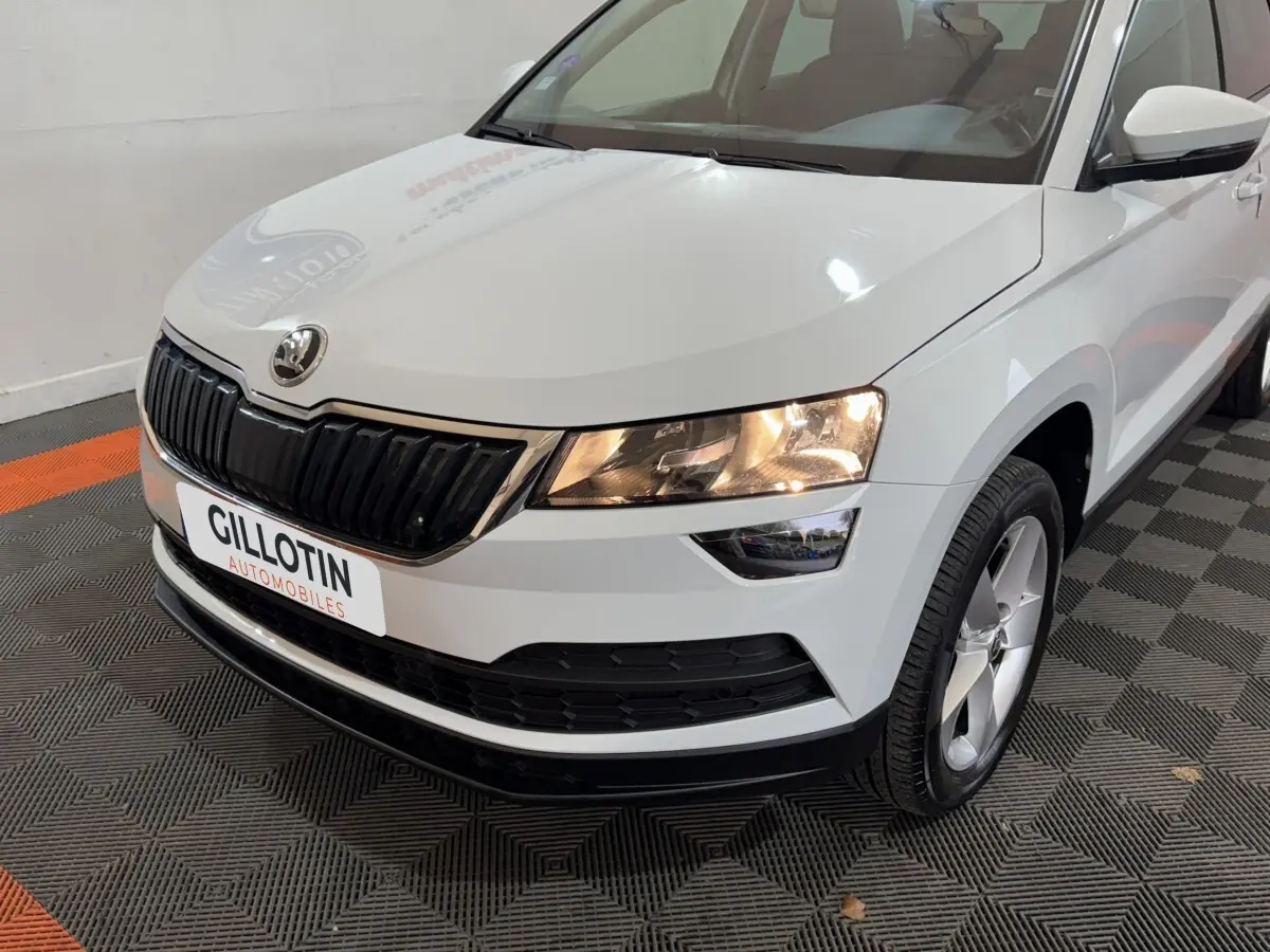 Vue 3/4 avant droite d'un Skoda Karoq blanc avec calandre noire et phares allumés en intérieur showroom.