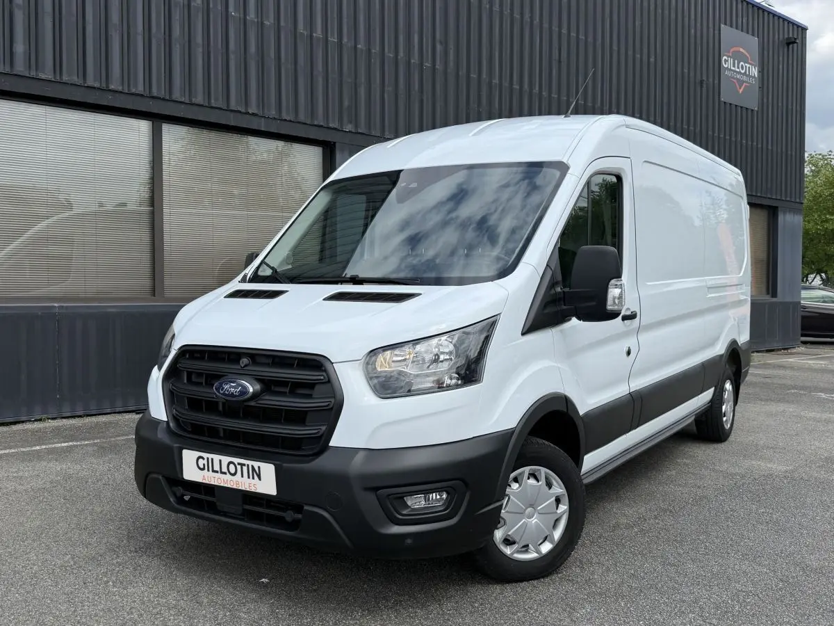 Ford Transit Fourgon blanc vu en 3/4 avant droit, avec calandre noire et feux allumés devant un bâtiment industriel.