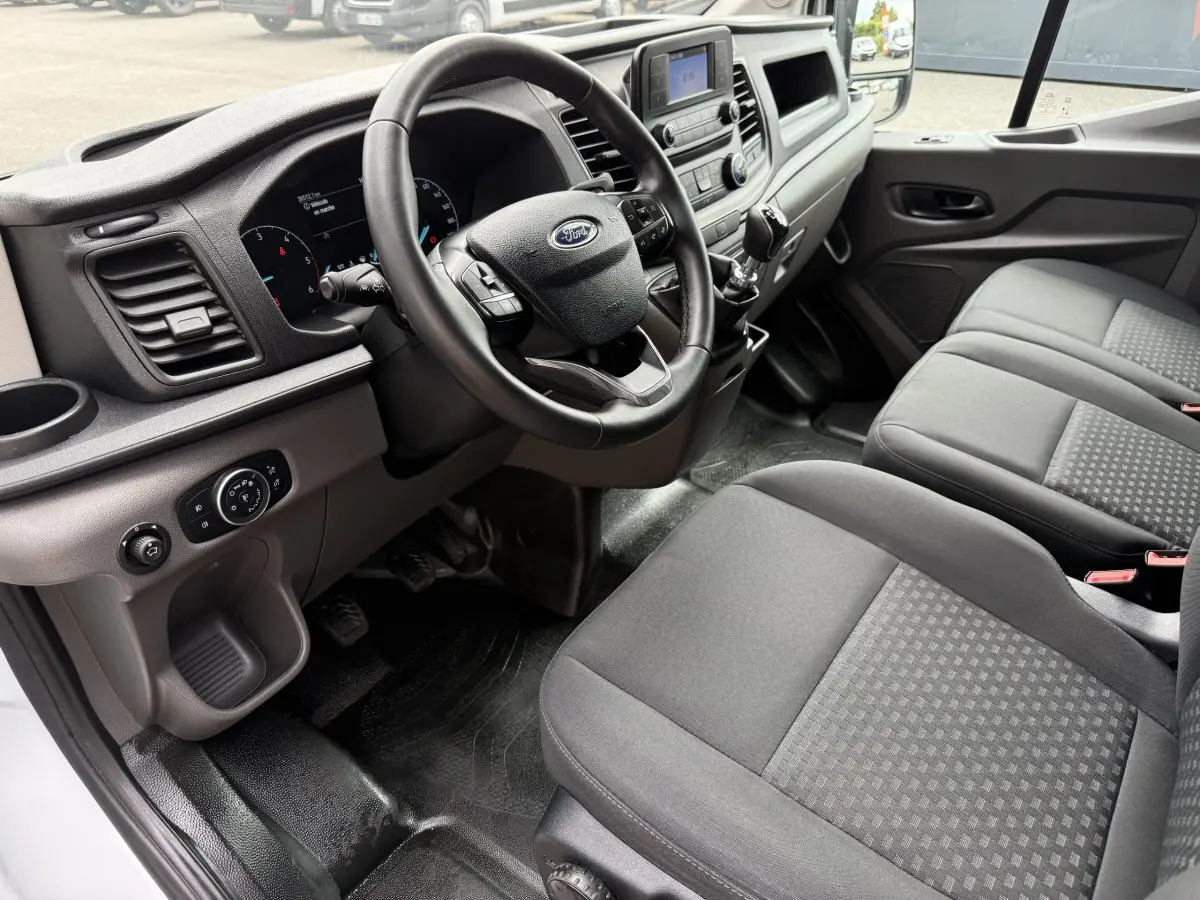 Vue intérieure côté conducteur du Ford Transit Fourgon 2024, montrant le volant cuir, tableau de bord et sièges gris.
