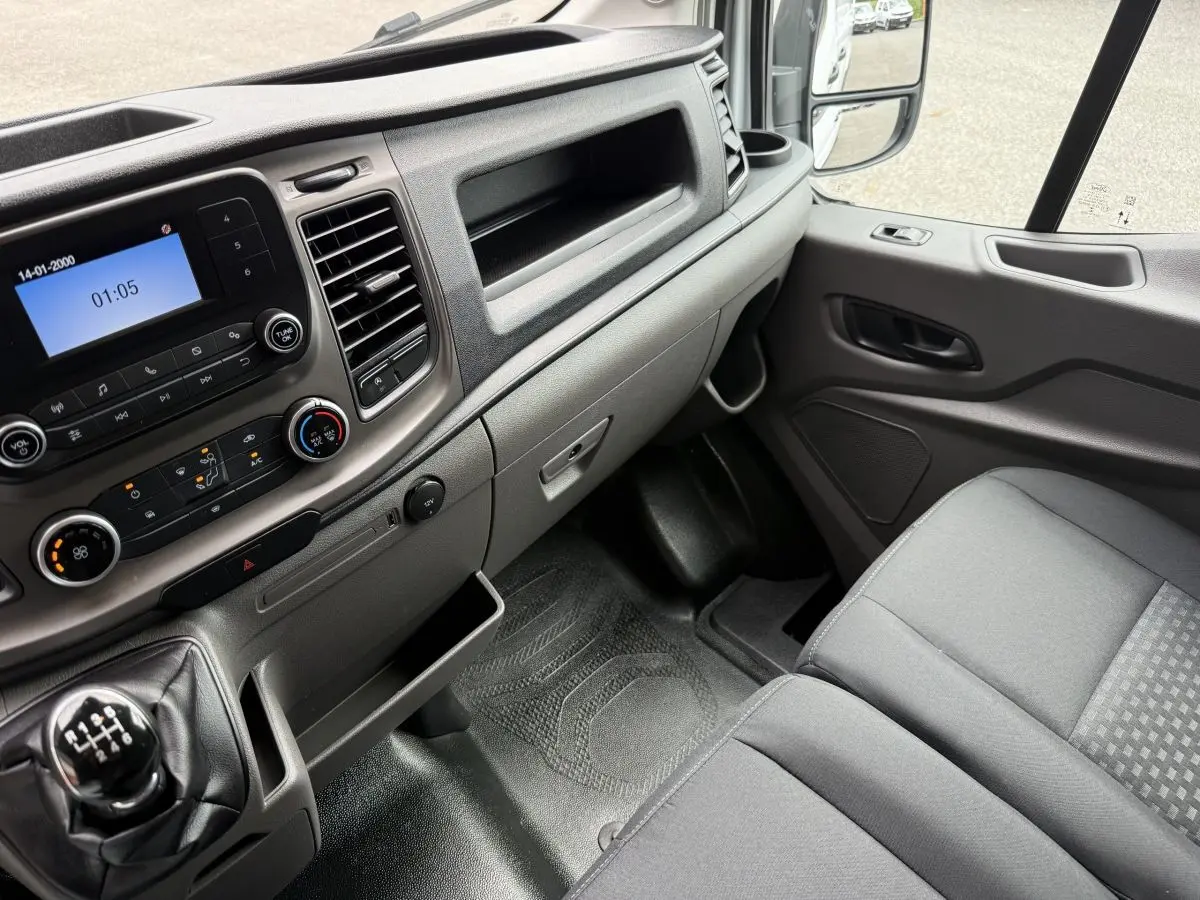 Vue intérieure côté passager du Ford Transit Fourgon 2024, tableau de bord gris et siège tissu gris.