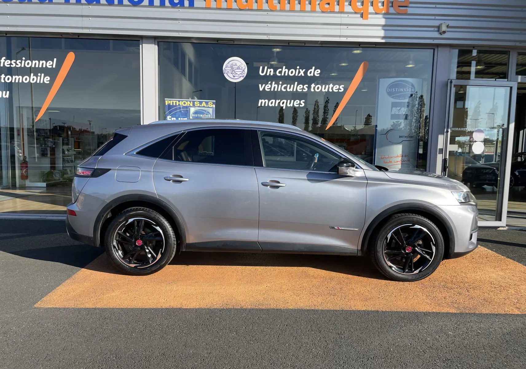 Profil côté gauche d'un DS7 gris Artens 2018 avec jantes noires, garé devant une vitrine de concession multimarque.