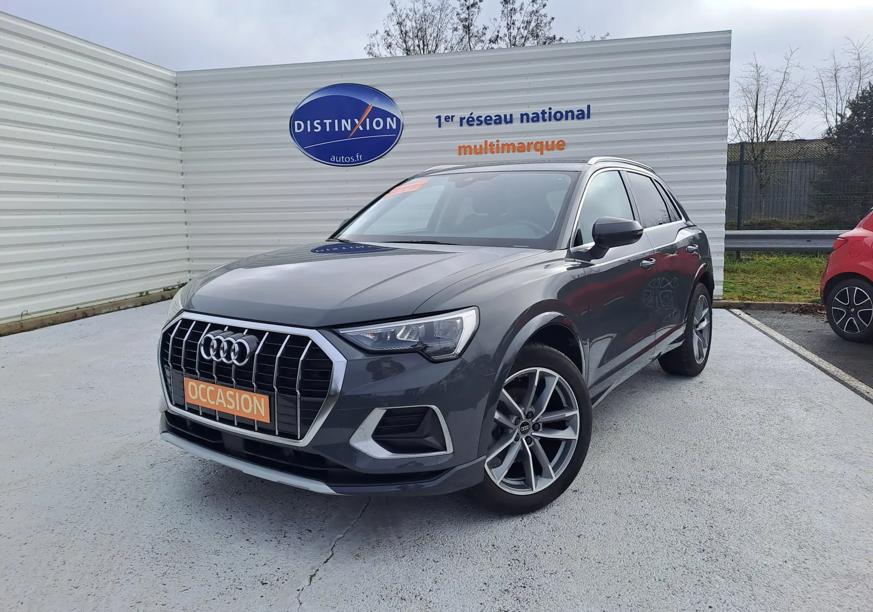 Audi Q3 gris nano en 3/4 avant droit, avec jantes alu 19 pouces et calandre chromée visible sur parking extérieur.