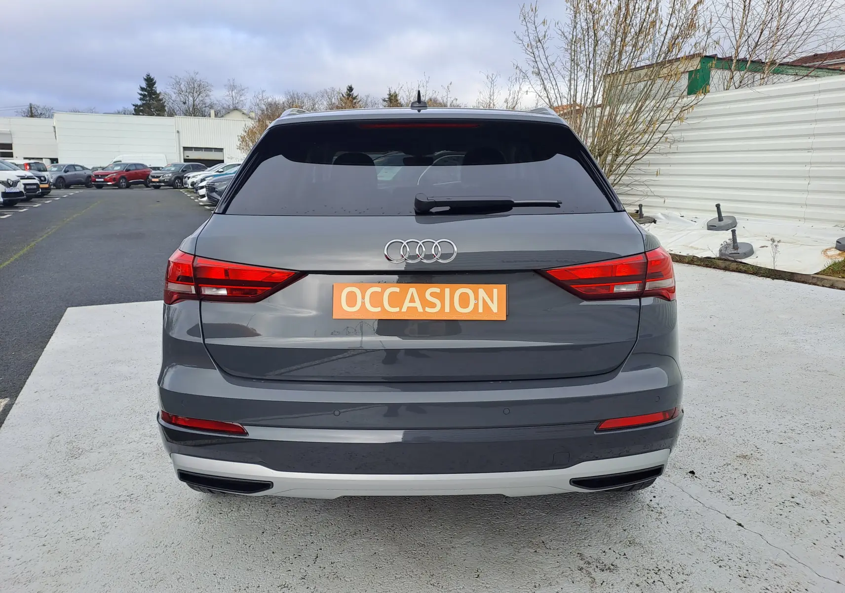 Vue arrière d'un Audi Q3 gris nano avec feux LED et plaque orange "OCCASION" sur un parking extérieur.