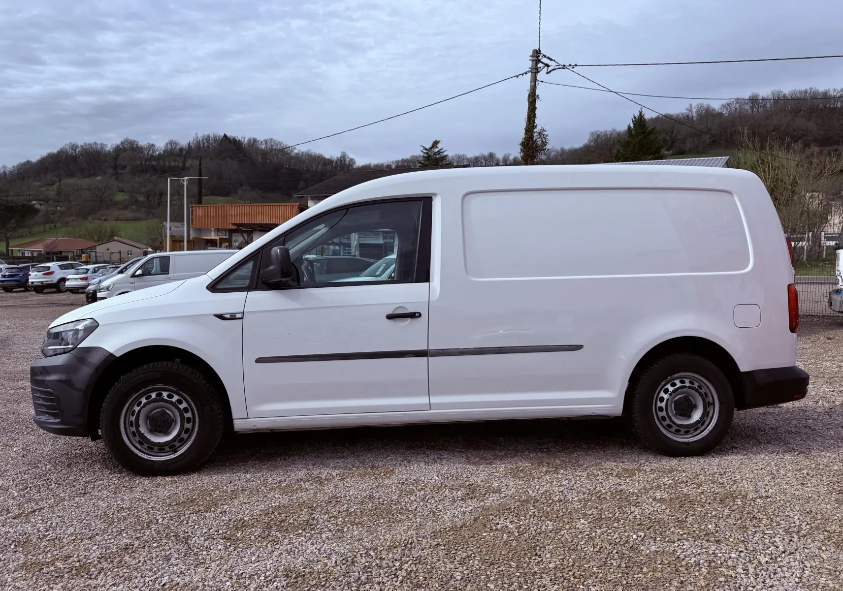 Vue de profil côté gauche d’un Volkswagen Caddy Maxi Long blanc utilitaire avec pare-chocs noirs sur un terrain gravillonné.