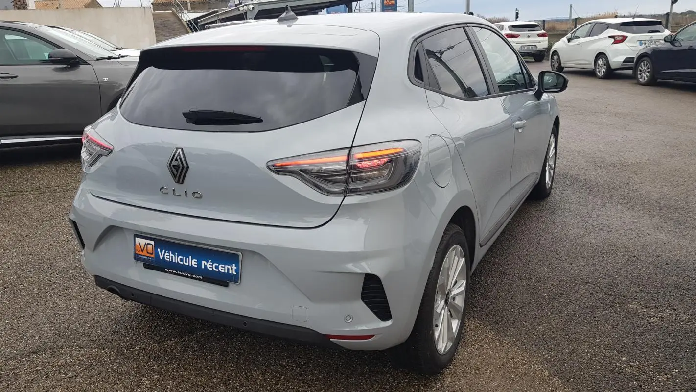 Vue 3/4 arrière droite d'une Renault Clio gris clair avec vitres surteintées et feux arrière allumés.