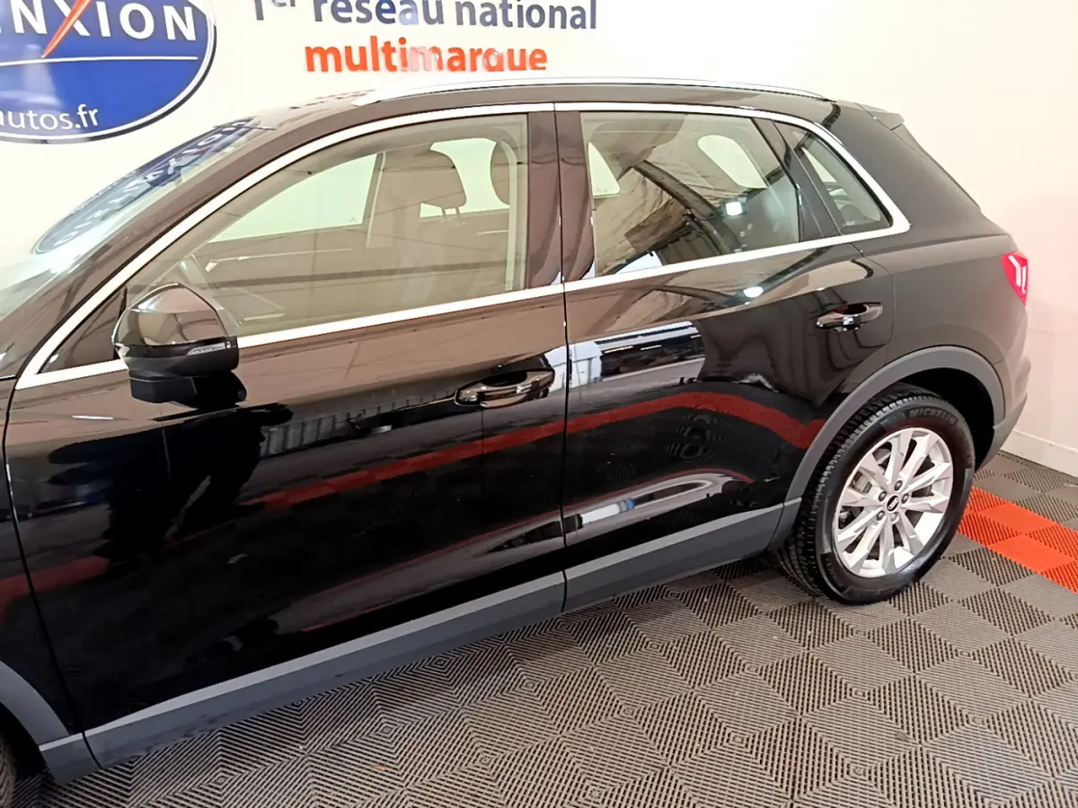 Profil côté gauche noir brillant de l'Audi Q3 35 TDI 150 S Tronic Business Line avec jantes argentées en intérieur showroom.