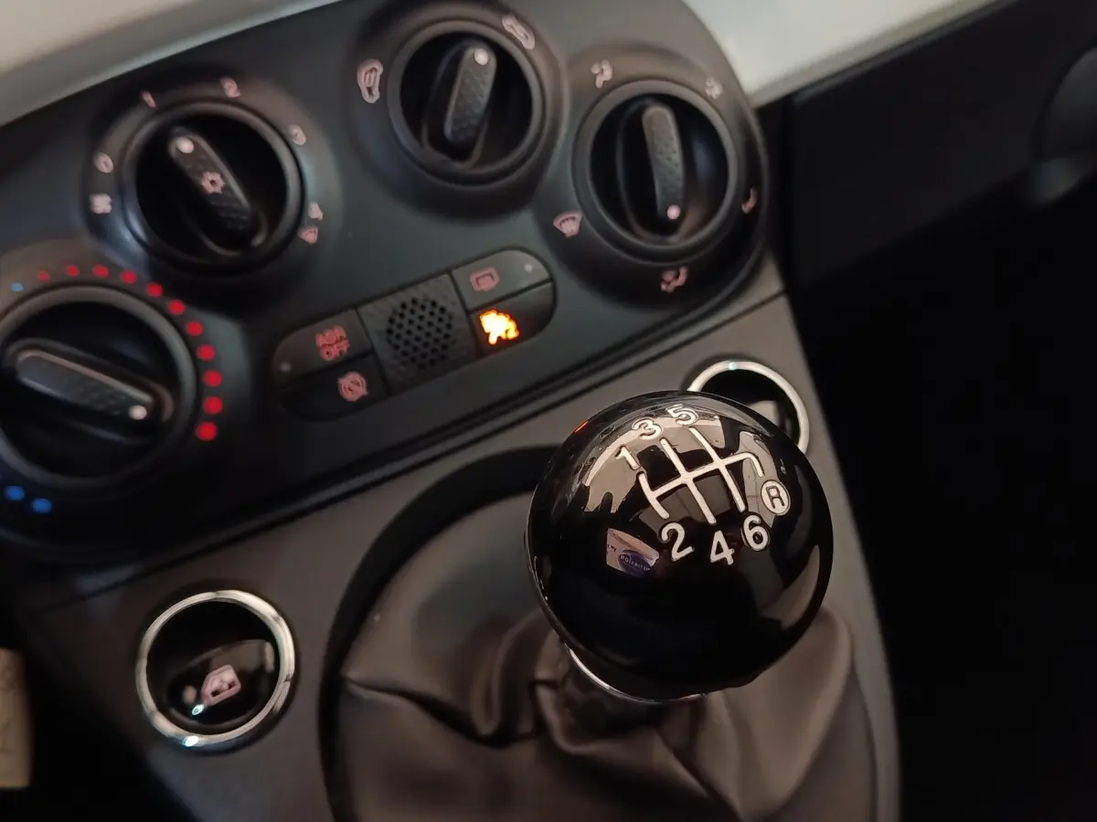 Gros plan sur le levier de vitesses manuel noir et le tableau de bord avec commandes de climatisation d'une Fiat 500 blanche.