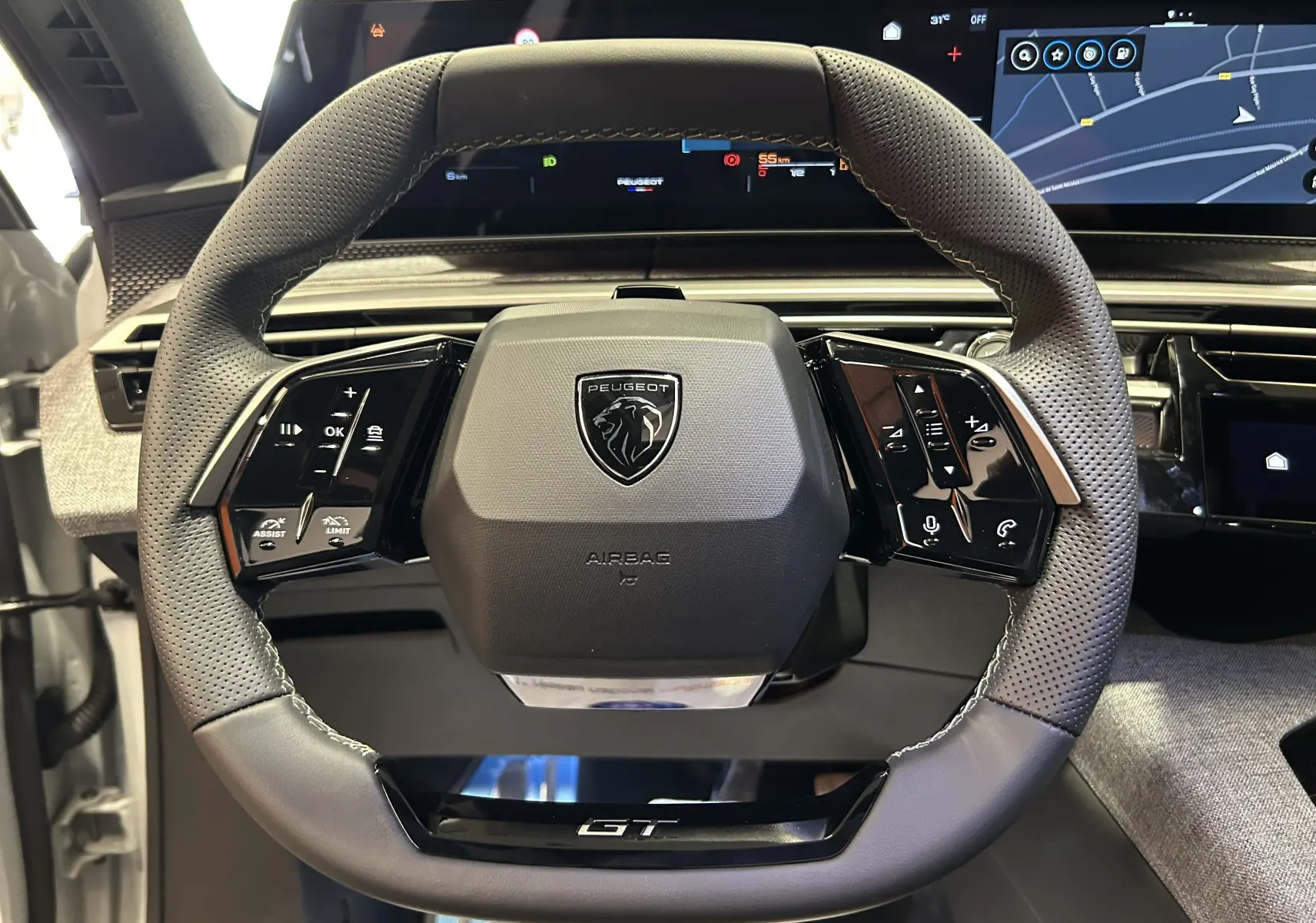 Vue rapprochée du volant GT perforé du Peugeot 3008 Hybrid 2026 avec commandes intégrées et écran numérique en arrière-plan.