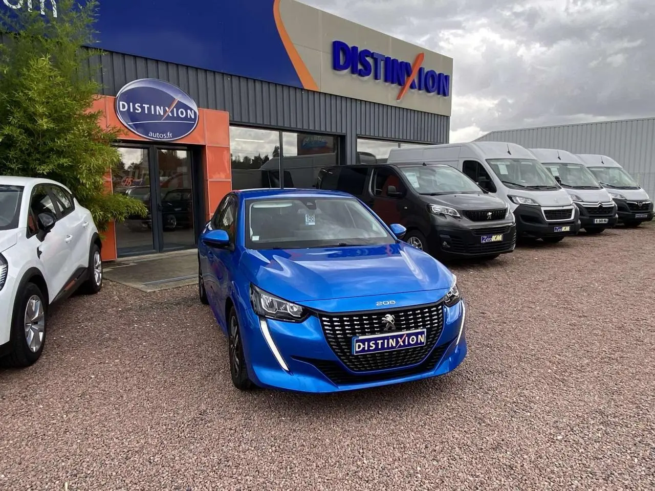 Peugeot 208 bleu en 3/4 avant stationnée devant un concessionnaire avec plusieurs véhicules utilitaires en arrière-plan.