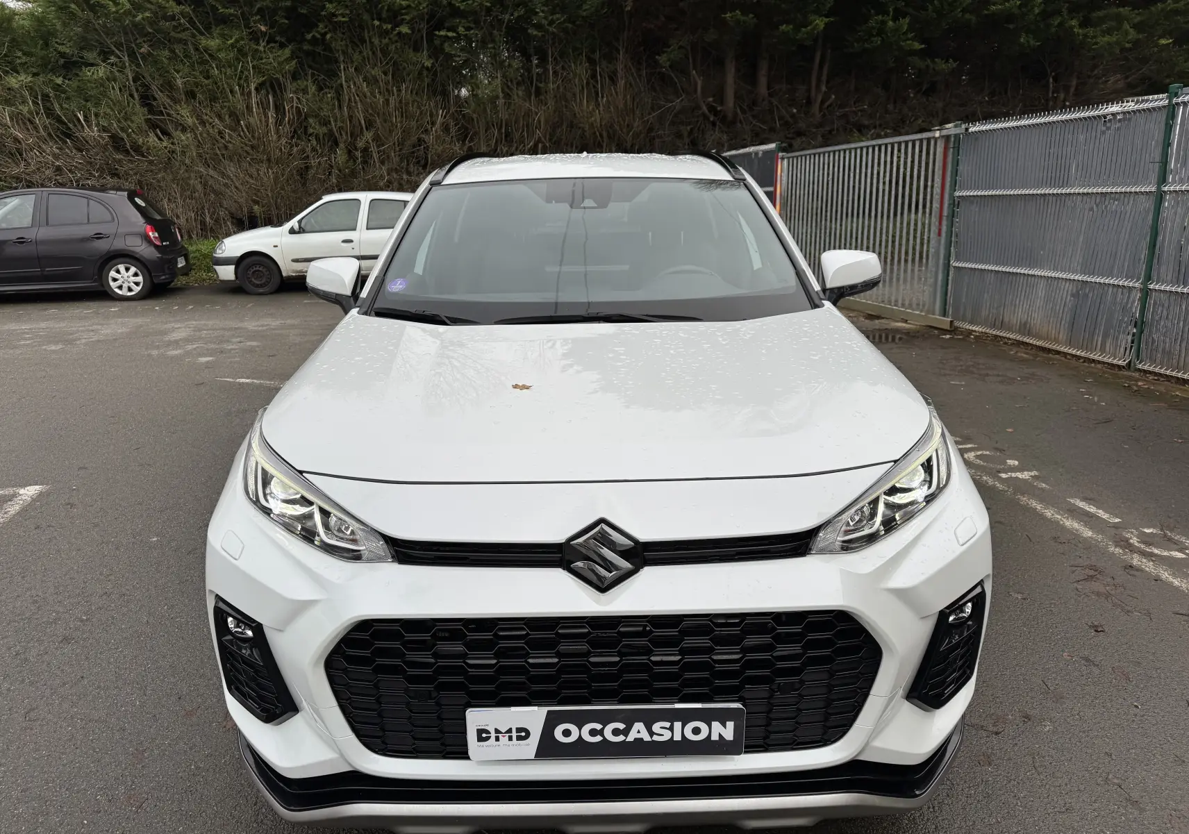 Vue frontale d'un Suzuki Across blanc 2023 avec calandre noire et phares LED, stationné en extérieur sur parking.
