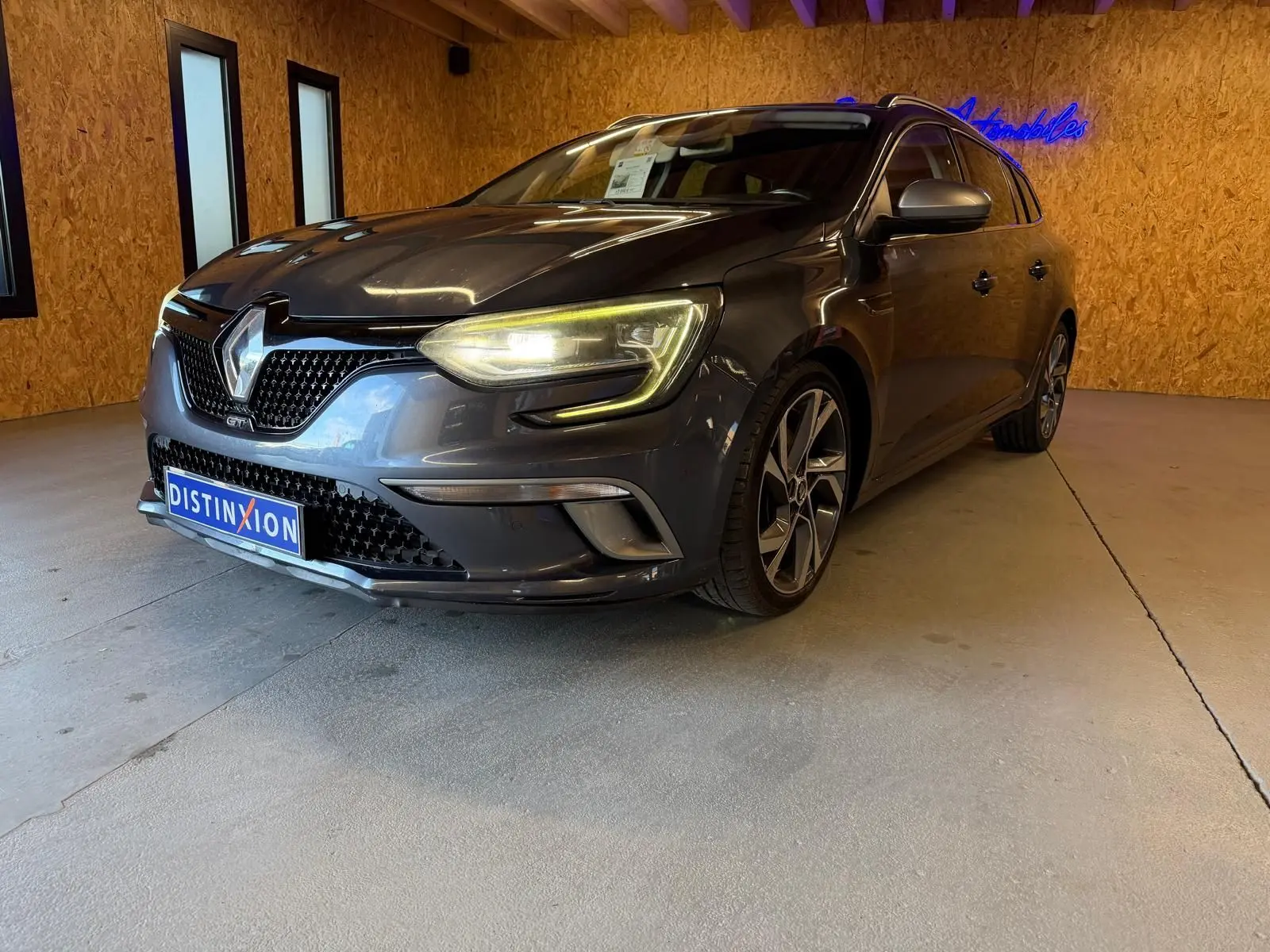 Renault Mégane Break GT gris vue 3/4 avant droit, phares allumés, dans un intérieur avec murs en bois.