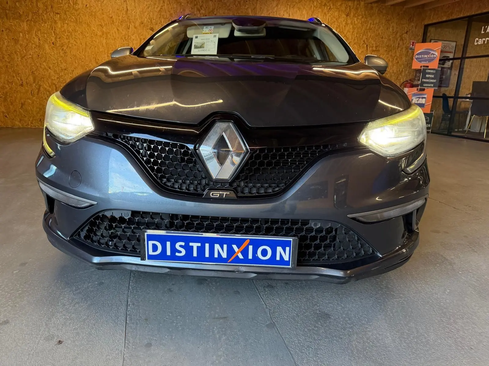 Vue de face d'une Renault Mégane Break GT gris avec phares allumés et calandre noire en intérieur showroom.