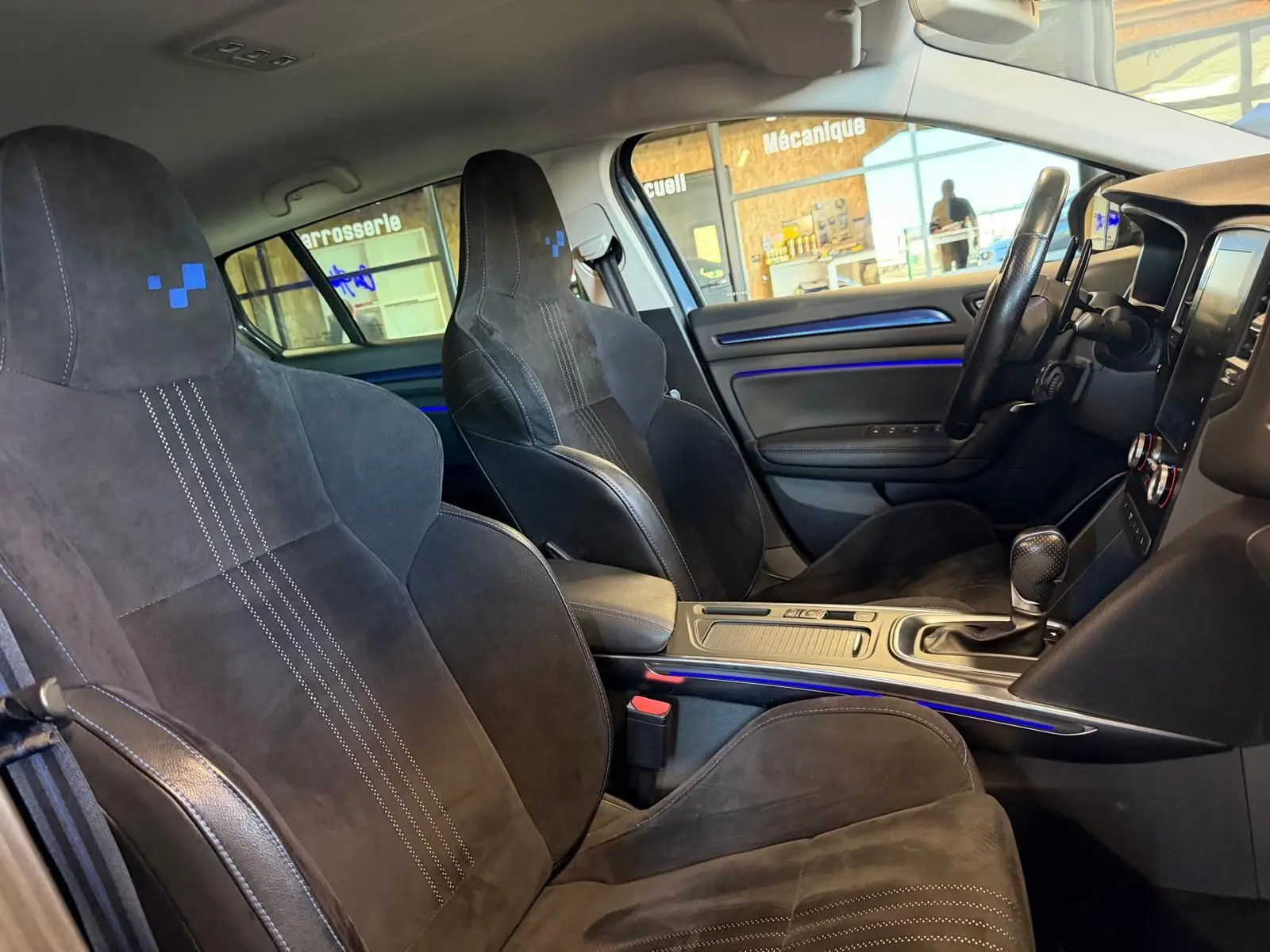 Vue intérieure côté conducteur de la Renault Mégane Break GT 2017, sièges sport noirs avec surpiqûres blanches et éclairage d'ambiance bleu.