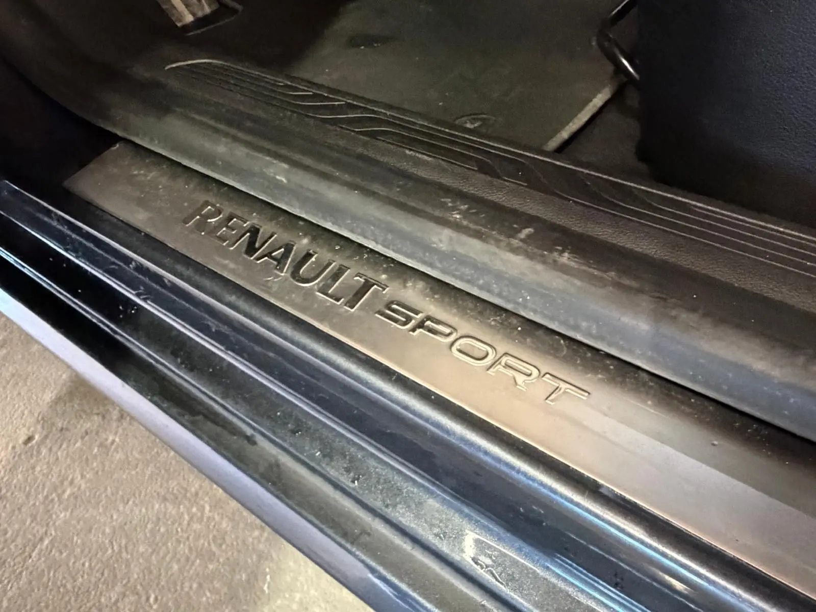 Gros plan sur la plaque de seuil de porte "RENAULT SPORT" noire, intérieur sombre du véhicule visible.