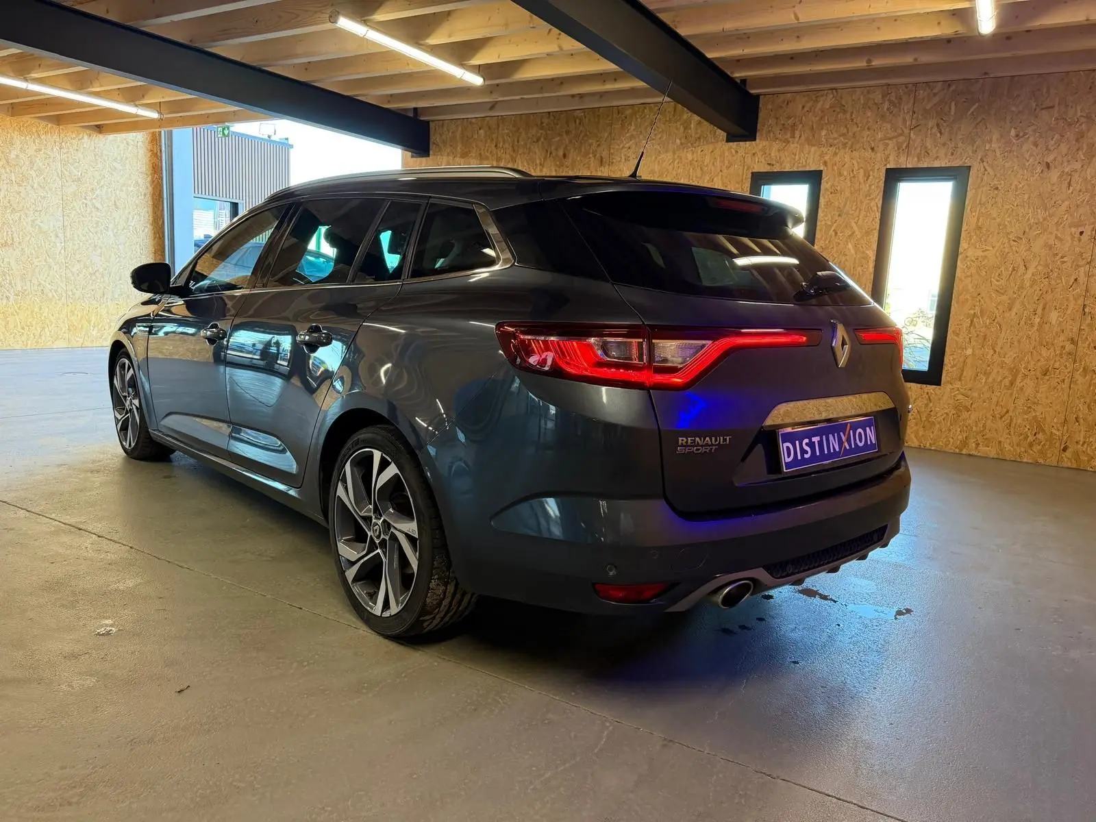Renault Mégane Break GT gris vue 3/4 arrière droit en intérieur, feux arrière allumés et jantes sport visibles.