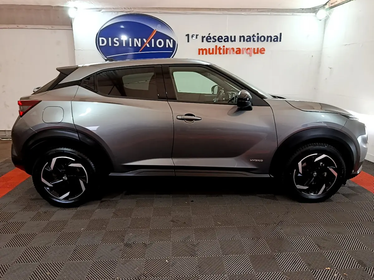 Profil côté droit d'un Nissan Juke Hybrid 143 Business+ gris foncé avec jantes noires et logo hybride visible sur l'aile avant.