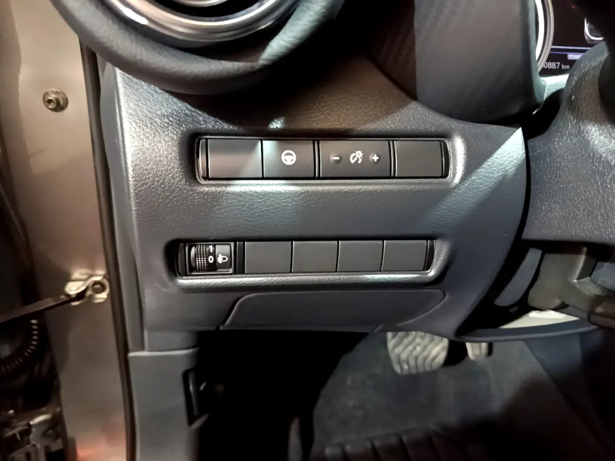 Gros plan sur les commandes de réglage de luminosité et assistance au volant côté gauche du tableau de bord du Nissan Juke gris foncé.